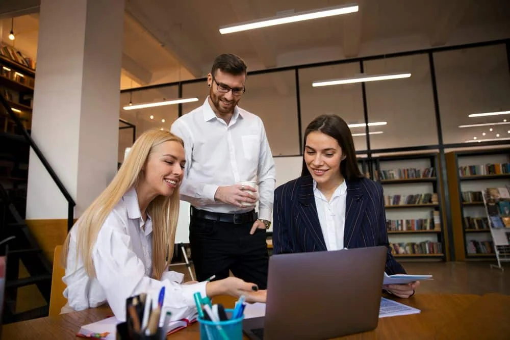 res personas jóvenes en un entorno de oficina moderna colaboran alrededor de un computador portátil. Dos mujeres están sentadas, una señalando la pantalla mientras la otra sostiene documentos; un hombre de pie las acompaña sonriendo con una taza en la mano. La escena refleja trabajo en equipo y ambiente profesional amigable, con estanterías de libros al fondo.